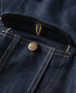 Indigo denim jacket detail: dark inner yoke and gold-tone brand stamp on pocket