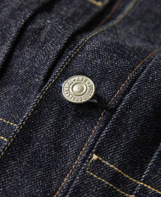Indigo denim jacket pocket flap with metal button and contrast gold stitching.