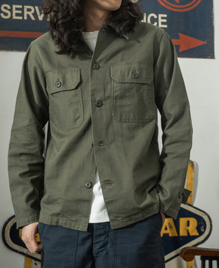 Olive fatigue utility shirt worn open over a T-shirt, chest pockets visible
