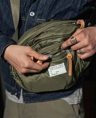 Olive waist bag with front zip pocket and hanging label, shown while worn