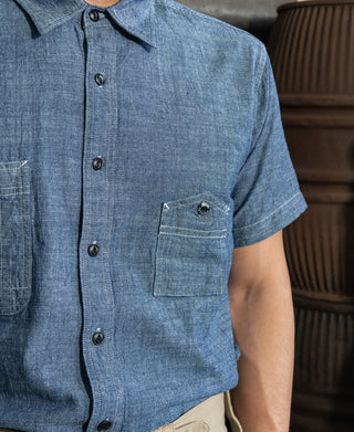 Blue chambray shirt front with pointed collar, black buttons, and chest pocket.