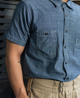 Blue chambray work shirt with short sleeves, chest pocket, and black button placket.