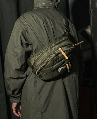 Olive green waist bag worn in front, textured quilted body with orange zipper tabs