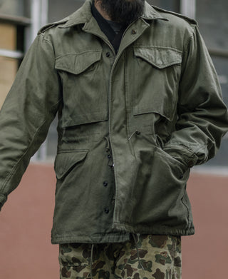 Olive field jacket with flap chest pockets and snap front, worn over camo pants