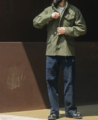 Olive field jacket with button front and chest flap pockets worn with dark pants.