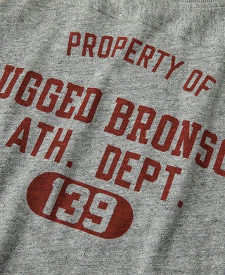 Gray T-shirt chest graphic: red “PROPERTY OF RUGGED BRONSON ATH. DEPT. 139”