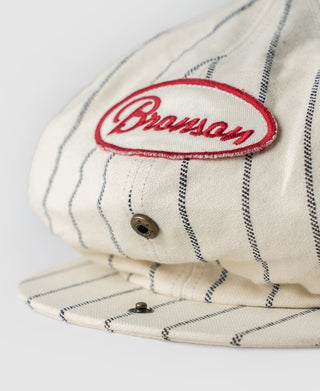 White pinstripe newsboy cap close-up with Bronson logo, button, and brim fold