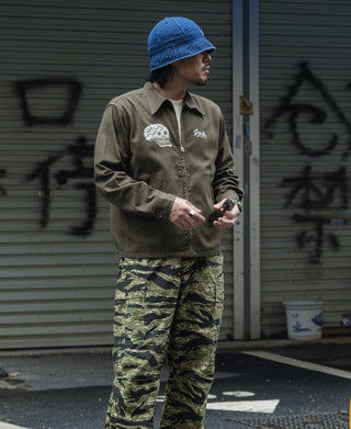 Olive zip-up souvenir jacket with skull chest embroidery and blue bucket hat styling