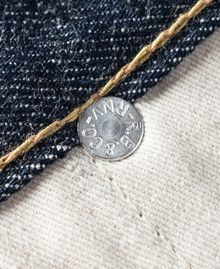 Denim jeans selvedge edge with round metal rivet on white fabric backing.