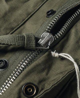 Olive field jacket close-up of two-way zipper, snap strap, and grommet hardware