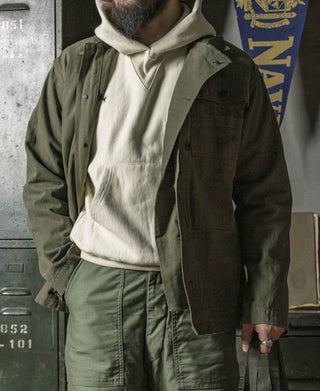 Olive utility jacket with button front and chest flap pockets, worn over hoodie