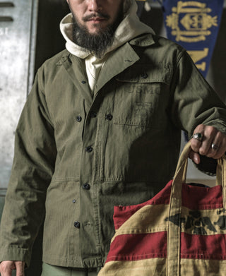 Olive utility jacket with dual chest flap pockets and button front, holding a bag