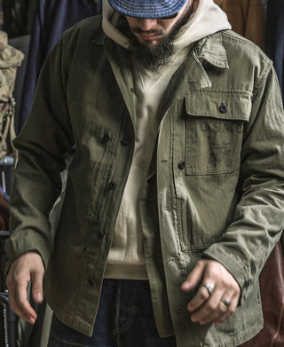 Olive utility jacket with flap chest pockets and button front, worn with hoodie and pants