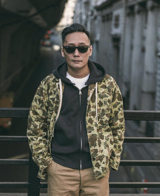 Camouflage duck hunter camo jacket worn over dark hoodie, front zip visible
