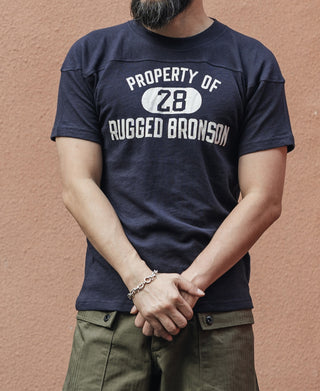 Model wearing navy t-shirt with front graphic “PROPERTY OF 28” and “RUGGED BRONSON” text
