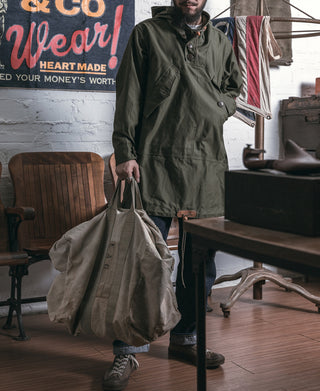 Olive hooded cotton parka with front buttons and drawstring, holding a large tote bag