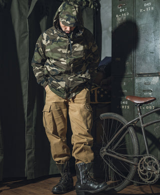 Woodland camo parka with hood up and front zipper, worn with boots.