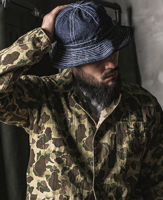 Indigo denim bucket hat with curved brim and stitched trim, worn with a camouflage jacket