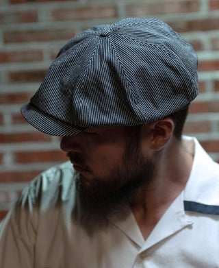 Striped denim newsboy cap side view in dark blue with white pinstripes and curved brim.